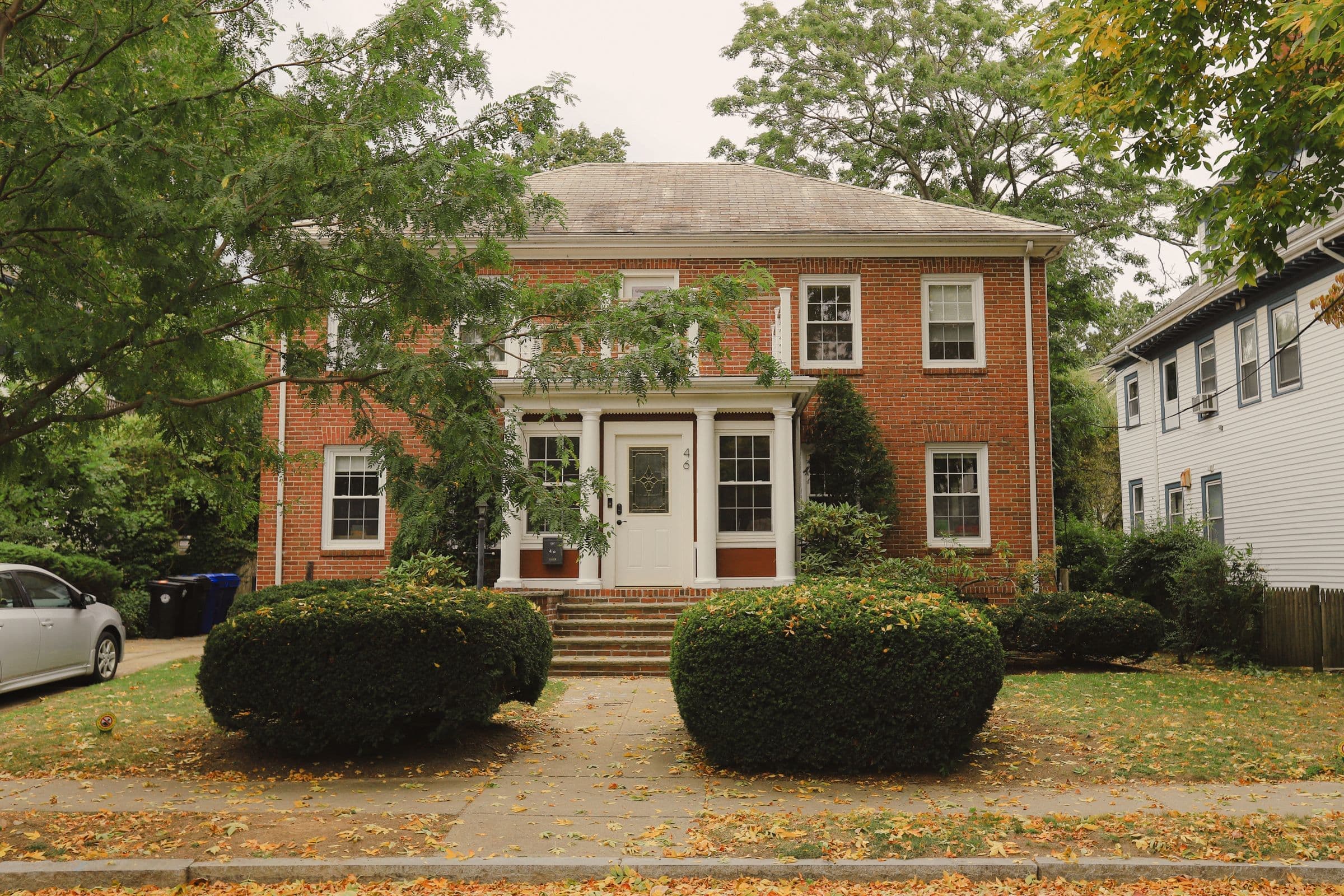 Junk removal in Alcoa, TN: older two-story brick house behind trimmed shrubs on a tree-lined street with fallen leaves