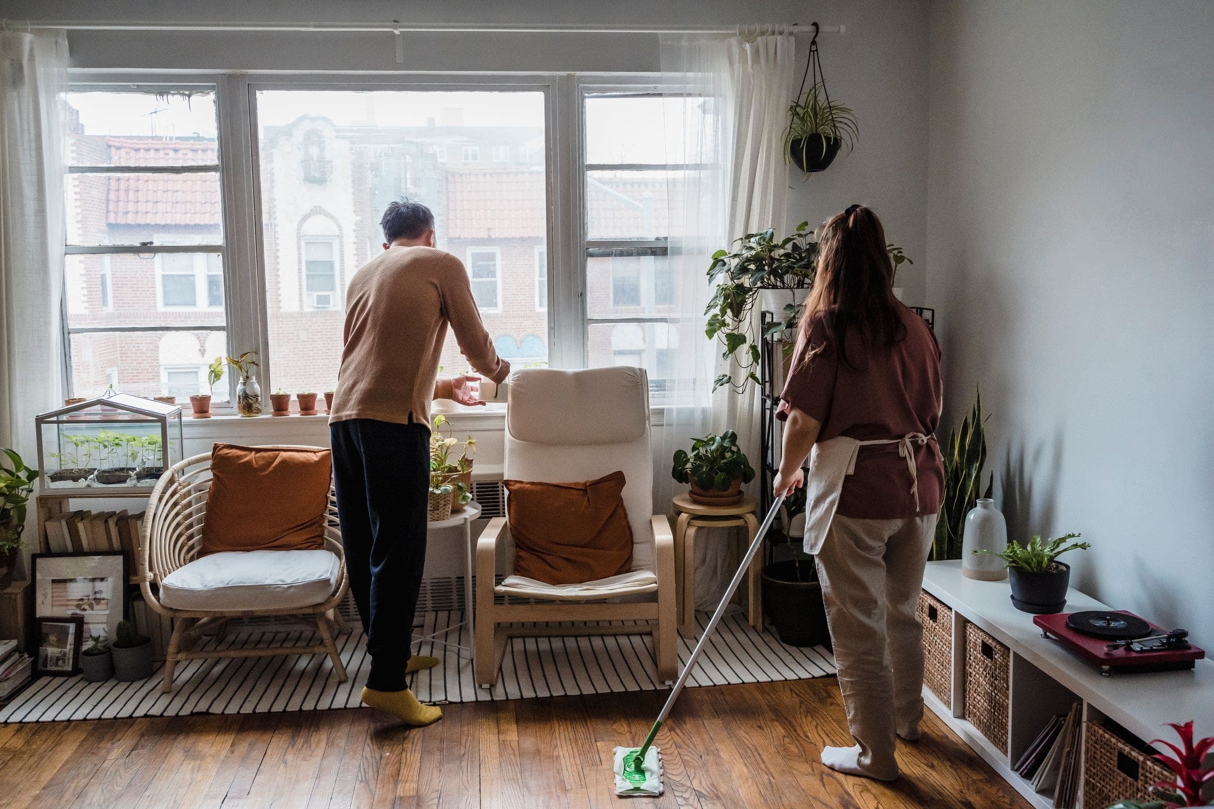 A couple tidying their living room, one mopping the wood floor and one tending plants by the window